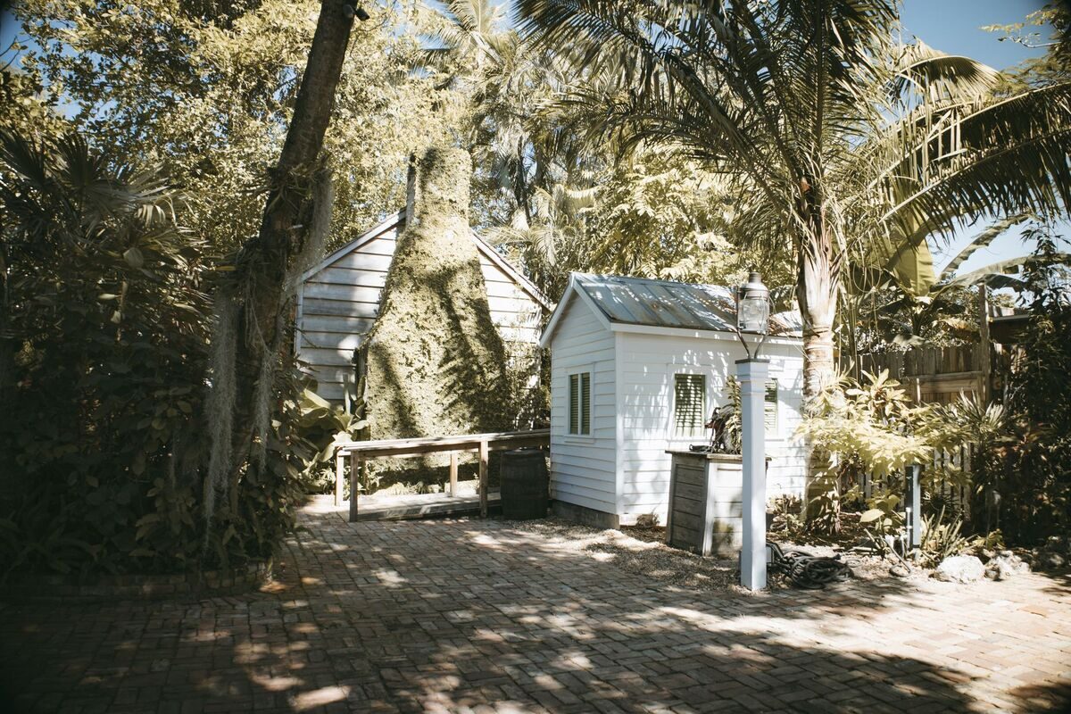 White garden shed beneath fan palms and lush subtropical plants on a brick patio in dappled Florida light