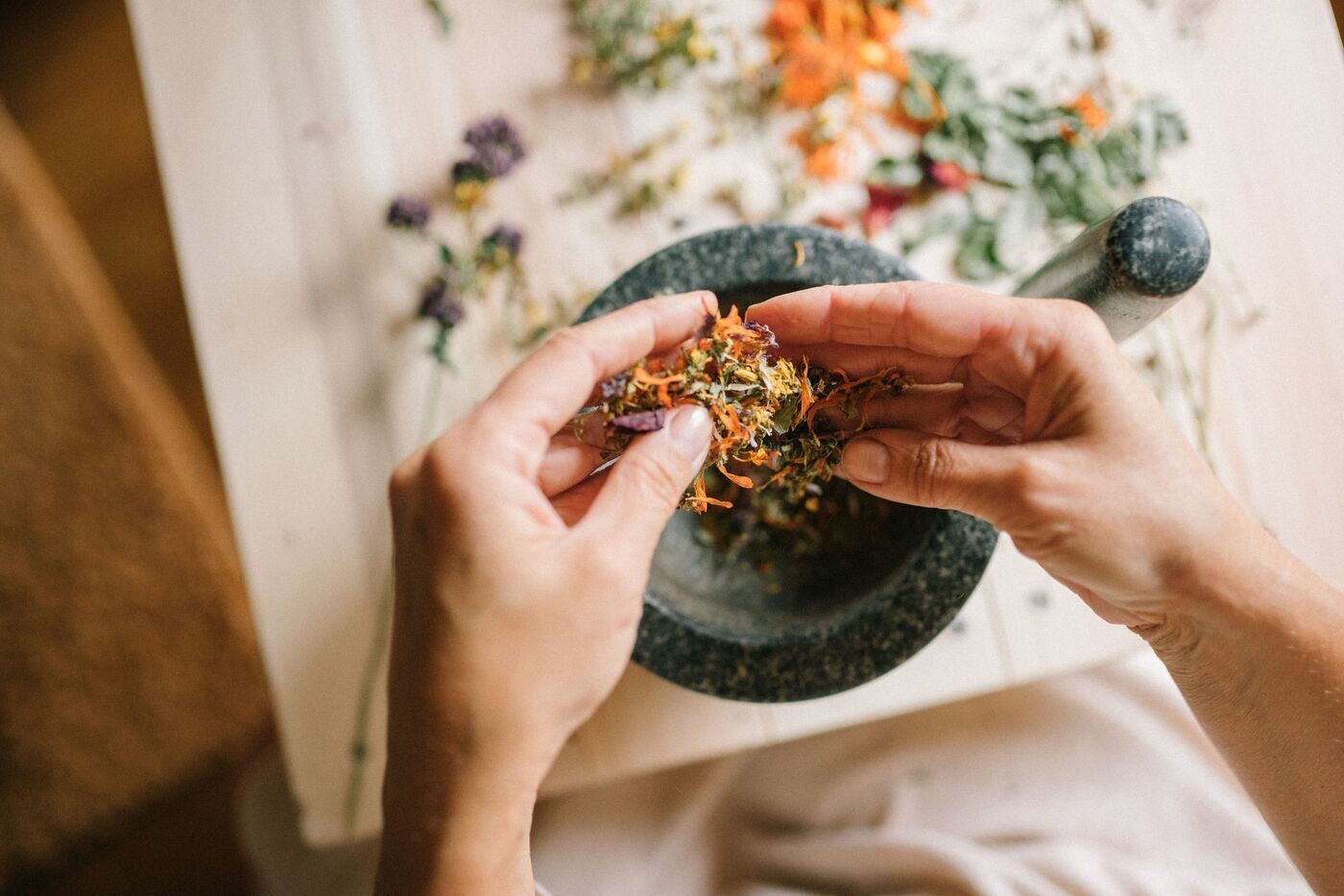 Hands crumbling colorful dried flowers and herbs over a stone mortar