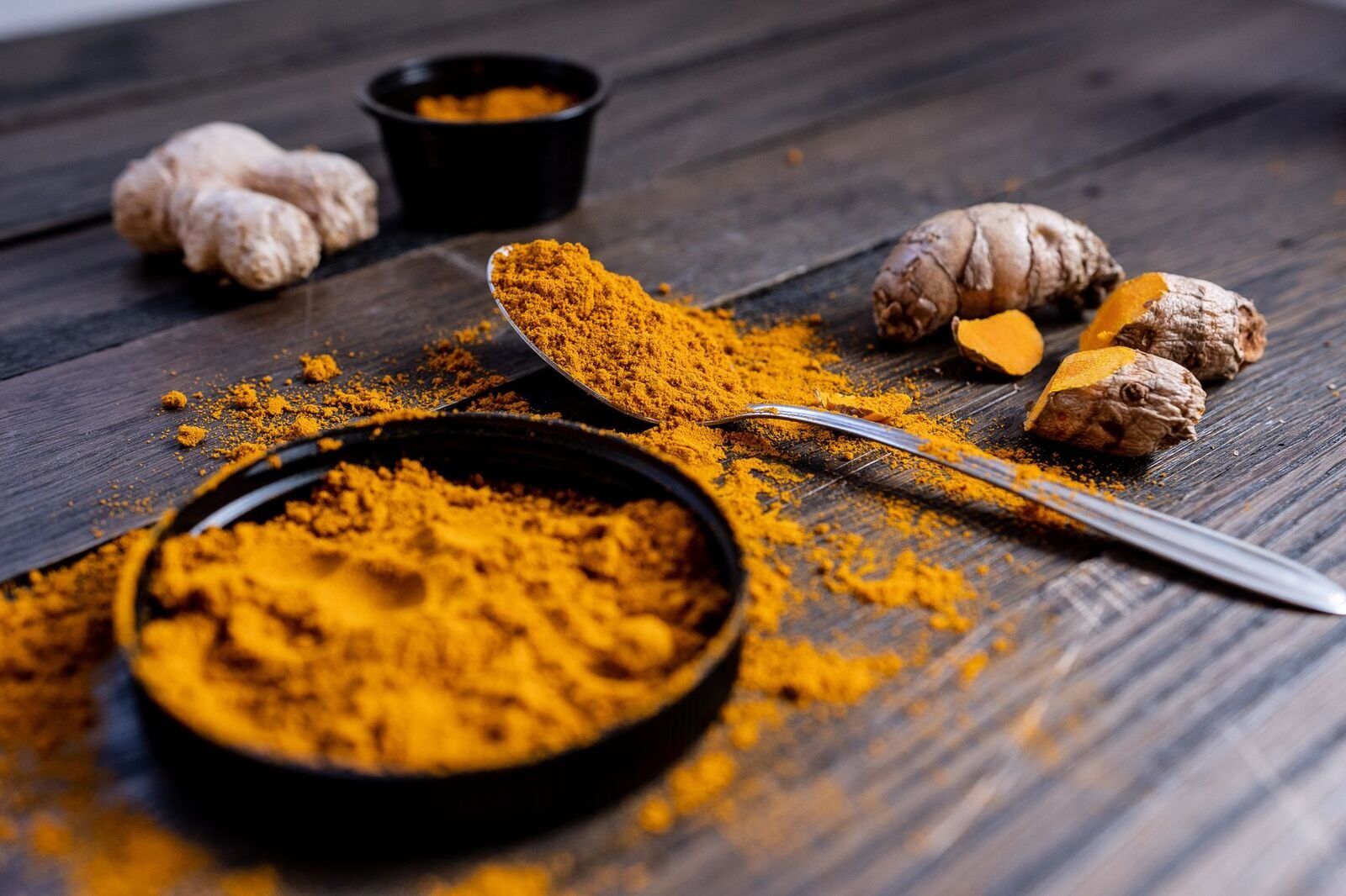 Golden turmeric powder spilled across a spoon and dark wooden table beside fresh turmeric roots