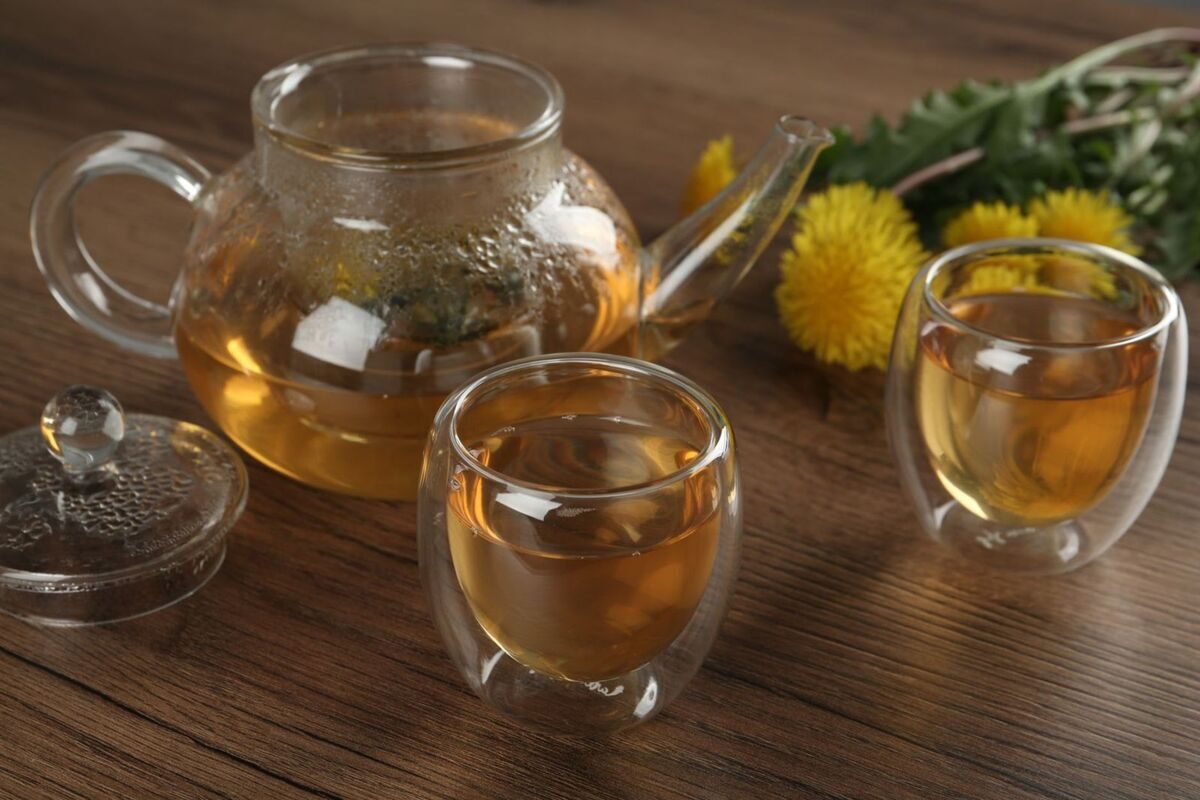 Glass teapot and cups of amber herbal infusion beside fresh dandelion flowers on a wooden table