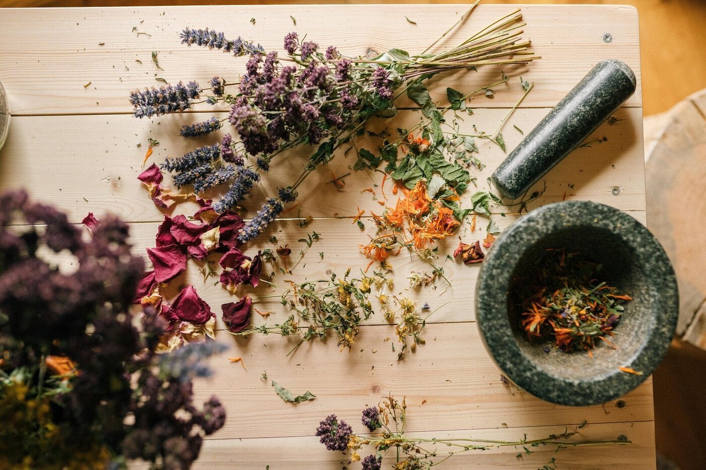 Dried lavender, rose petals, and calendula scattered on pale wood beside a stone mortar and pestle