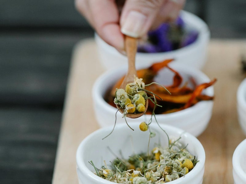 A hand spooning dried chamomile flowers over small bowls of botanicals